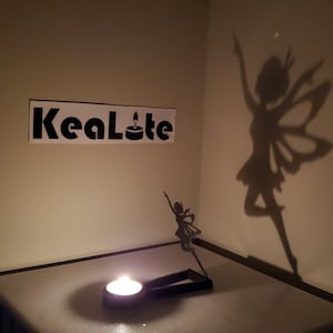May include: A black metal fairy silhouette candle holder with a tea light candle. The candle holder casts a shadow of a fairy on the wall behind it. The text "RealLite" is visible on the wall.