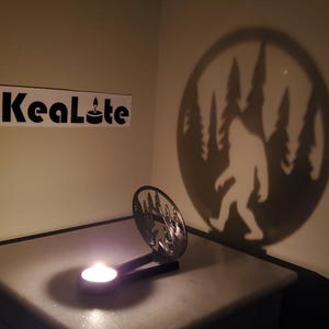 May include: A metal candle holder with a silhouette of Bigfoot walking through a forest. The candle holder casts a shadow on the wall behind it, showing the silhouette of Bigfoot. The candle holder is on a white surface. The text "KeaLote" is visible on a white sticker on the wall.