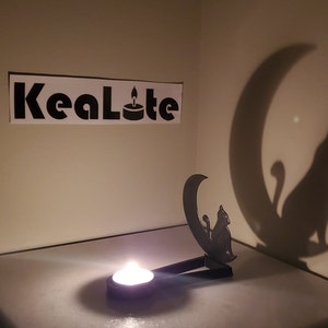 May include: Black metal candle holder shaped like a cat sitting on a crescent moon. The candle holder casts a shadow of a cat on the wall. The candle holder is lit with a tea light candle. The text "KeaLote" is visible on the wall.