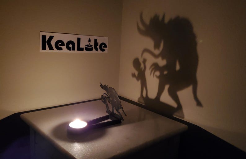 May include: A metal silhouette of a monster and a child casting a shadow on a wall. The silhouette is lit by a candle in a tealight holder. The text "KeaLite" is visible on a white sticker on the wall.