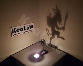 May include: A candle holder casting a shadow of a demon on a wall. The shadow is large and detailed, with horns and a tail. The candle holder is on a white surface, and the text "KeaLite" is visible.