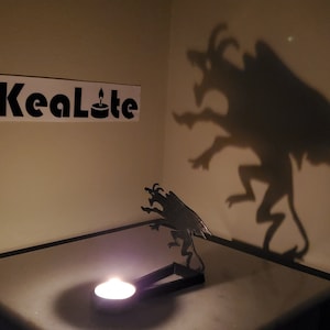 May include: A black metal candle holder shaped like a three-headed dragon with a lit tea light candle inside. The dragon casts a shadow on the wall behind it.