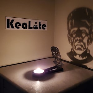 May include: A black metal candle holder shaped like Frankenstein's monster casts a shadow on the wall. The candle holder is lit with a tea light candle. The text "KeaLite" is visible on the wall.