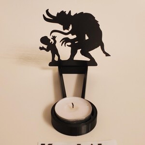 May include: Black metal candle holder featuring a silhouette of a demon and a child. The holder has a round base for a tea light candle.