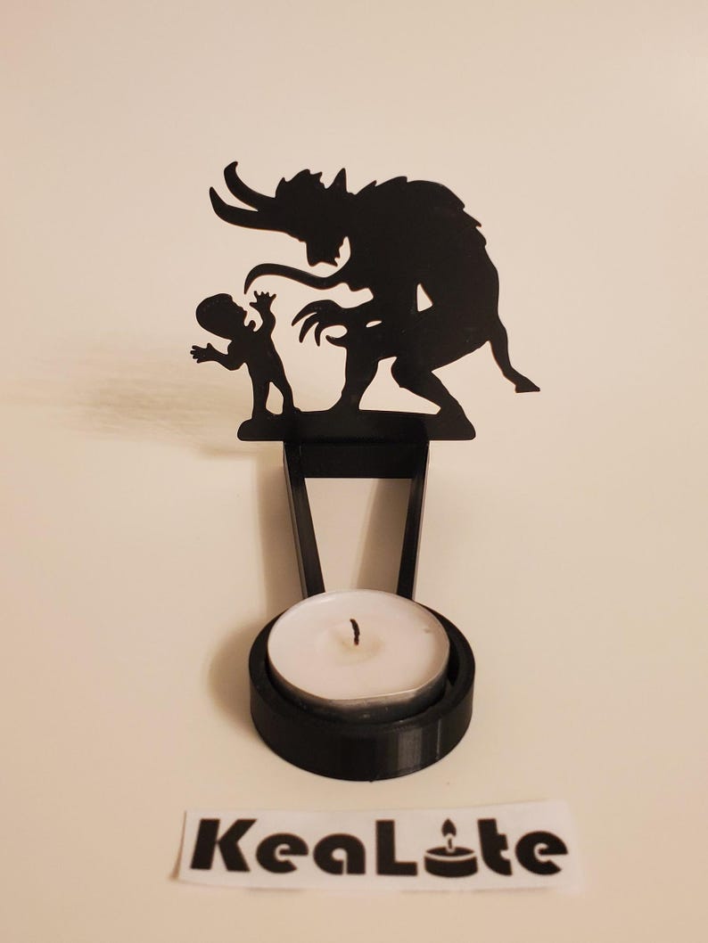 May include: Black metal candle holder featuring a silhouette of a demon and a child. The holder has a round base for a tea light candle.
