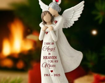 May include: A white angel ornament with a red ribbon for hanging. The ornament depicts two figures embracing, with one figure having wings. Text on the ornament reads "I HAVE AN Angel IN HEAVEN I CALL HER Mom 1961 - 2025".