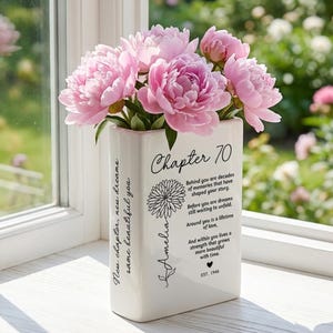 May include: A white rectangular ceramic vase filled with pink peonies. The front features the text Chapter 70, a flower illustration, the name Amelia, and a poem about memories and dreams. The side displays the text New chapter, new dreams, same beautiful you.