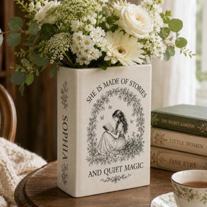 May include: A rectangular cream-colored vase with the text "SHE IS MADE OF STORIES AND QUIET MAGIC" and the name "SOPHIA". The vase is decorated with a floral illustration of a woman reading. The vase holds a bouquet of white flowers.