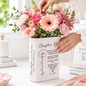 May include: A white rectangular ceramic vase featuring black printed text that reads Chapter 40, a floral illustration, the name Amelia, and a poem about memories and dreams. The vase is filled with a vibrant bouquet of pink roses, gerbera daisies, and greenery.
