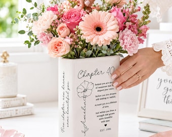 May include: A white rectangular ceramic vase featuring black printed text that reads Chapter 40, a floral illustration, the name Amelia, and a poem about memories and dreams. The vase is filled with a vibrant bouquet of pink roses, gerbera daisies, and greenery.