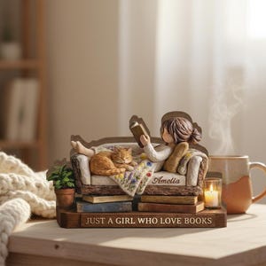 May include: A figurine of a girl reading on a couch with a sleeping cat. The couch is atop a stack of books, with the text "JUST A GIRL WHO LOVE BOOKS" on the base. A small plant, candle, and mug are also present.