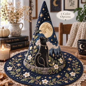 May include: A quilted navy blue witch hat featuring an embroidered black cat sitting on a stone wall, a large full moon, white flowers, and gold stars. The hat displays a silhouette of a castle and includes text stating 4 Color Options.