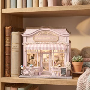 May include: A miniature pink and white cat cafe with the text "Neko & Pastel Cafe" above the entrance. The cafe features a striped awning, a bicycle with a basket, and several cat figurines inside. A small chalkboard sign reads "Cat Pudding & Paw-stries".