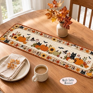 May include: A rectangular quilted table runner featuring a festive autumn and Halloween theme with orange pumpkins, sunflowers, black cats, ghosts wearing witch hats, and small bats on a cream background, bordered by a brown and tan plaid fabric.