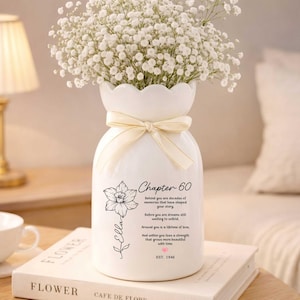 May include: A white ceramic vase with a scalloped rim and a cream-colored ribbon tied around the neck. The vase features a black floral line drawing, the name Ella, the text Chapter 60, a poem, a small pink heart, and the text EST. 1946.