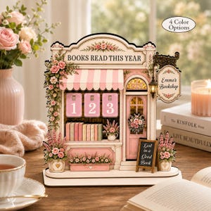 May include: A decorative wooden book nook kit designed as a miniature bookstore named Emma's Bookshop. It features a pink and white striped awning, floral accents, a sign reading BOOKS READ THIS YEAR, and a chalkboard sign saying Lost in a Good Book.