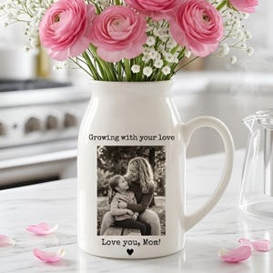 May include: A white ceramic vase with a handle, filled with pink flowers and white filler flowers. The vase has a black and white photo of a mother and child, with the text "Growing with your love" and "Love you, Mom!"