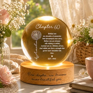 May include: A spherical glass ornament illuminated on a wooden base. The glass features the text 'Chapter 60', a floral graphic, the name 'Amelia', a poem about memories and dreams, and 'EST. 1966'. The wooden base is engraved with 'New chapter, new dreams same beautiful you'.