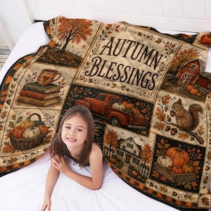 May include: A quilted throw blanket featuring a patchwork design with autumn-themed illustrations including a red truck, pumpkins, a squirrel, a house, and books. The text AUTUMN BLESSINGS is printed in the center in a dark brown serif font.