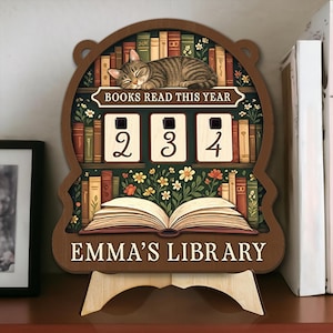 May include: Wooden bookshelf-shaped sign with a sleeping cat, floral accents, and the text "BOOKS READ THIS YEAR." It features a counter displaying "234" and the words "EMMA'S LIBRARY." The sign is supported by a wooden stand.