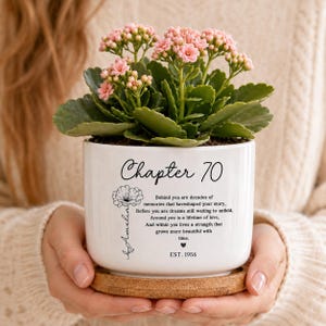 May include: A white ceramic planter with a bamboo saucer containing a pink Kalanchoe plant. The planter features the text Chapter 70, a floral graphic, the name Amelia, a poem about memories and strength, and the text EST. 1956 printed in black ink.