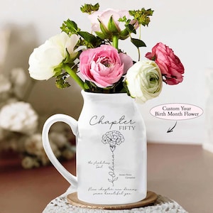May include: A white ceramic pitcher with a handle, filled with a bouquet of pink, white, and red flowers. The pitcher has the words "Chapter FIFTY" and a floral illustration. Text on the pitcher reads "Custom Your Birth Month Flower."