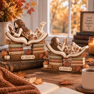 May include: Two decorative figurines of a girl with long brown hair lying on an open book, wearing a dress with autumn leaf patterns. The base features stacked books labeled Just A Girl Who Loves Books and a nameplate with the name Julia.