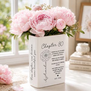 May include: A white ceramic rectangular vase filled with light pink peony flowers. The front features black text reading Chapter 80, a floral line drawing, the name Amelia, a poem, a heart icon, and EST. 1946. The side displays the text New chapter, new dreams, same beautiful you.
