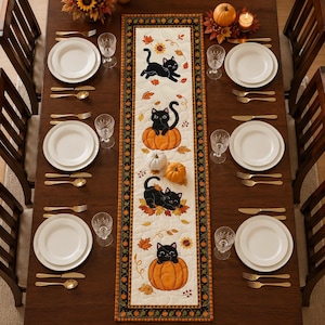 May include: A rectangular quilted table runner featuring a cream-colored center with illustrations of black cats and orange pumpkins, surrounded by a dark border with a repeating pumpkin pattern, set on a wooden dining table with white plates.