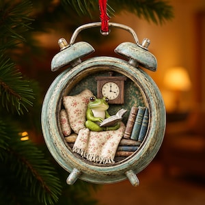 May include: A decorative hanging ornament shaped like a distressed teal alarm clock. Inside the clock face, a green frog sits on a floral patterned pillow and blanket, reading an open book. A small wall clock and a stack of books are visible.