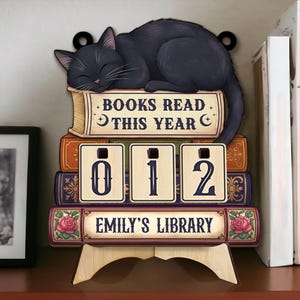 May include: Wooden bookshelf decor featuring a sleeping black cat atop a stack of books. The books display the text "BOOKS READ THIS YEAR" and "EMILY'S LIBRARY". The numbers "012" are displayed on the books. The decor is supported by a wooden stand.