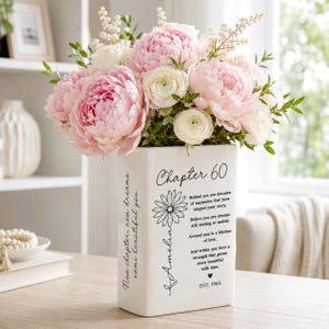 May include: A white rectangular ceramic vase filled with a bouquet of pink peonies and white ranunculus. The front features black text reading Chapter 60, a daisy illustration with the name Amelia, and a poem. The side reads New chapter, new dreams.