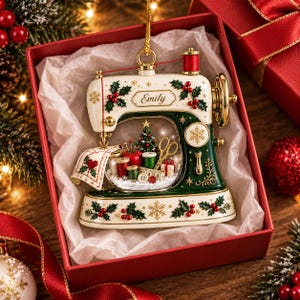 May include: A cream and dark green sewing machine ornament featuring the name Emily, decorated with holly, berries, and snowflakes. The center contains a miniature scene with a Christmas tree, scissors, thread spools, and a pincushion.