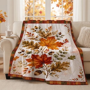 May include: A quilted throw blanket featuring a central design of vibrant orange, red, and brown autumn leaves and acorns on a white background, framed by a plaid border in shades of orange, red, yellow, and green, draped over a sofa.