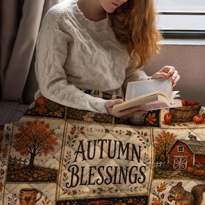 May include: A quilted throw blanket featuring a patchwork design with autumn-themed illustrations including trees, a red barn, pumpkins, and a squirrel. The central panel displays the text "AUTUMN BLESSINGS" surrounded by a decorative leaf border.