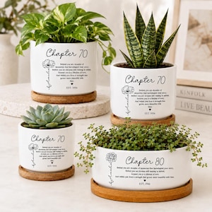 May include: Four white ceramic cylindrical planters with bamboo saucers, each featuring black text reading Chapter 70 or Chapter 80, a floral illustration, and a personalized name. The planters contain various green houseplants and succulents.
