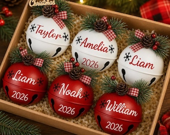 May include: A set of six round jingle bell ornaments in a cardboard box, three white and three red. Each ornament features a name, the year 2026, snowflake graphics, a pine sprig, a small pinecone, red berries, and a red and white checkered bow.