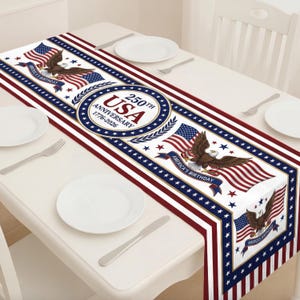 May include: A patriotic table runner featuring the American flag, an eagle, and the text "250th USA Anniversary 1776-2026." The design incorporates red, white, and blue colors, with stars and stripes. The runner is placed on a white table with plates and silverware.