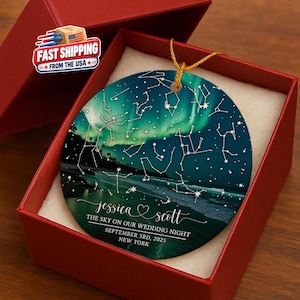 May include: A round ceramic ornament featuring a night sky with aurora borealis and star constellations. The text reads: Jessica & Scott, The sky on our wedding night, September 3rd, 2025, New York. It hangs by a gold string inside a red gift box.