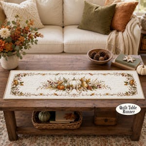 May include: A rectangular quilted table runner featuring a cream-colored fabric base with a printed design of autumn pumpkins, dried leaves, and berries. The border is decorated with a repeating pattern of fall foliage in shades of orange, brown, and green.