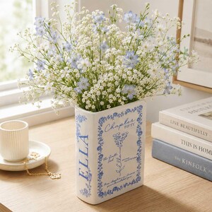 May include: A white ceramic book-shaped vase featuring blue floral borders and text that reads Chapter Fifty, Ella, and The Gentle Soul. The vase is filled with a bouquet of small white baby's breath and light blue delphinium flowers.