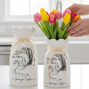 May include: Two white ceramic vases with scalloped tops and cream-colored ribbon. One vase holds colorful tulips. Each vase features a Winnie the Pooh illustration and the text "NEVER FORGET THE DIFFERENCE YOU MAKE. Thank you, Sophia."