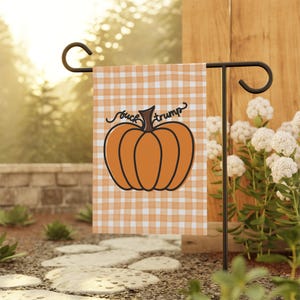 May include: A garden flag with an orange and white gingham pattern. The flag features an orange pumpkin graphic with the words "fuck trump" in black script. The flag is displayed on a black metal stand in a garden setting.