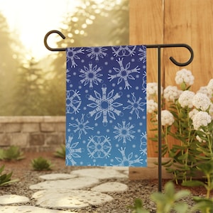 May include: A garden flag featuring a blue gradient background with white snowflake patterns. The flag is displayed on a black metal stand in an outdoor setting, with a stone pathway and greenery in the background.