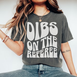 May include: A dark gray t-shirt with a white graphic that says "DIBS ON THE REFEREE" in a retro font.