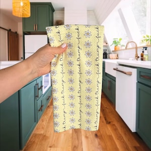 May include: A yellow kitchen towel with a daisy pattern and the text "fuck trump" in black script. The towel is held up in front of a kitchen with green cabinets and a white dishwasher.