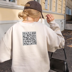 May include: A beige sweatshirt with a large black and white QR code printed on the back. The person is wearing a brown baseball cap and a black purse. The sweatshirt has long sleeves.