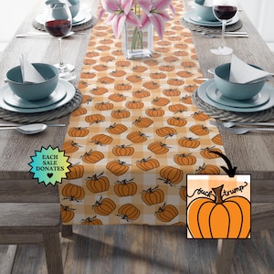 May include: A table runner with a repeating pattern of orange pumpkins on a white and orange checkered background. The runner is on a wooden table set for a meal, with plates, bowls, and wine glasses. The text "buck trump" is visible.