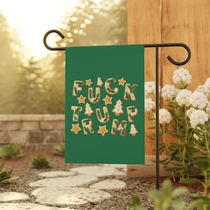 May include: A green garden flag with the phrase "FUCK TRUMP" spelled out in gingerbread cookie letters, stars, and Christmas tree shapes. The flag is displayed on a black metal stand in a garden setting.