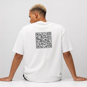 May include: White t-shirt with a large black QR code printed on the back. The shirt is a simple, casual style with short sleeves. The person in the image is wearing the shirt.